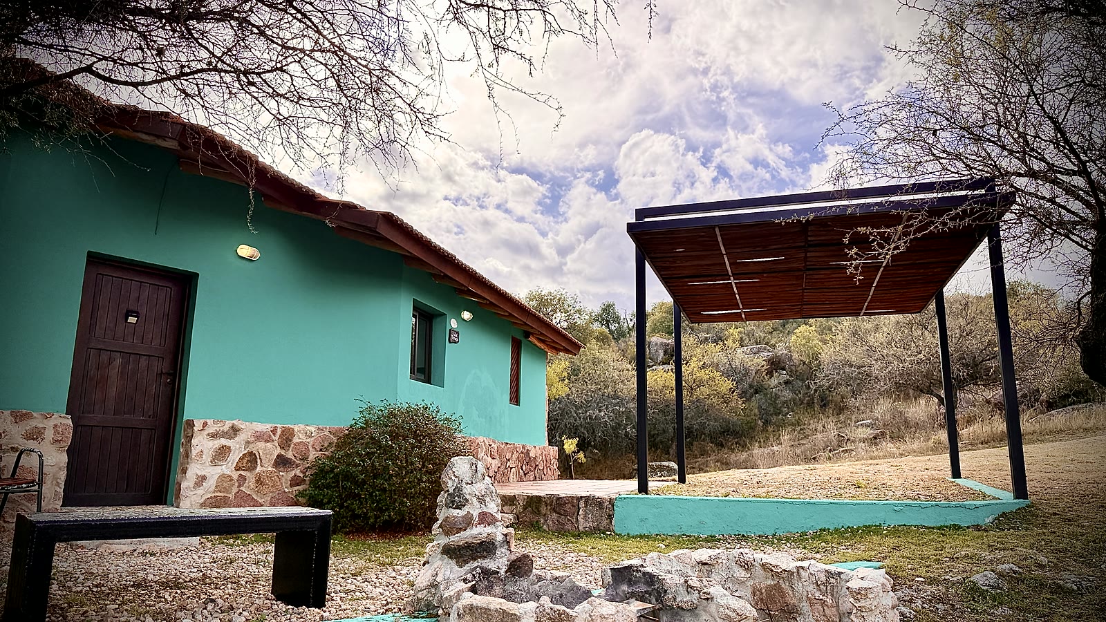 El frente turquesa de la cabaña, con la galería al costado y las sierras detrás